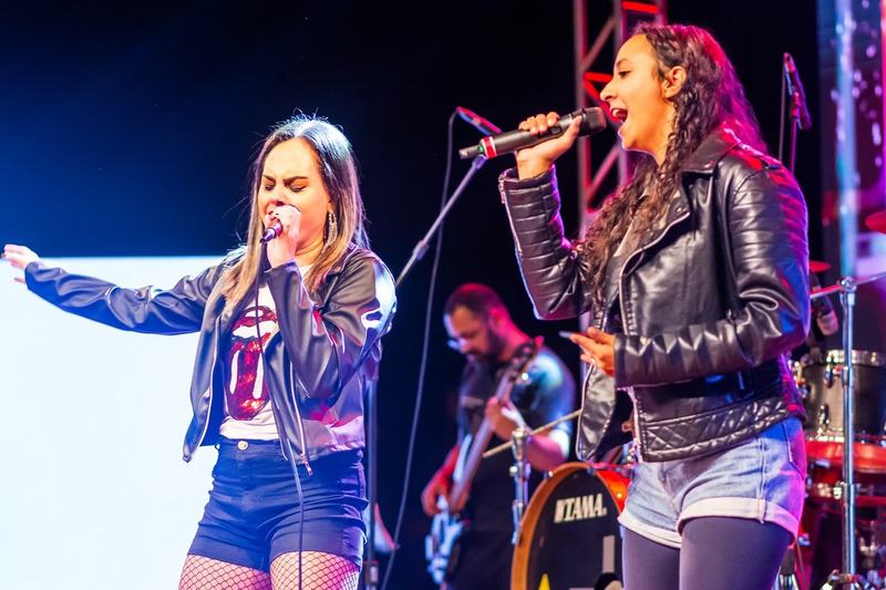 Duas cantoras de jaqueta de couro cantando no palco da apresentação, com o baixista atrás