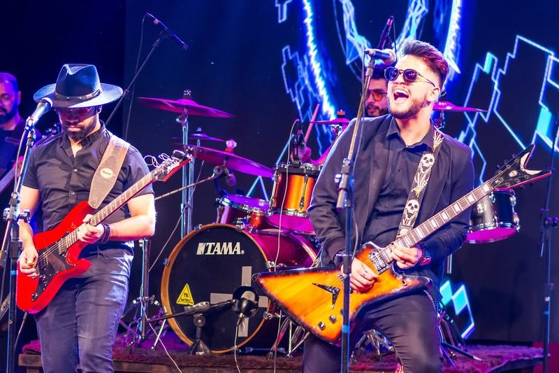 Dois guitarristas tocando numa apresentação, um de chapéu com guitarra vermelha e outro sorrindo com guitarra em V