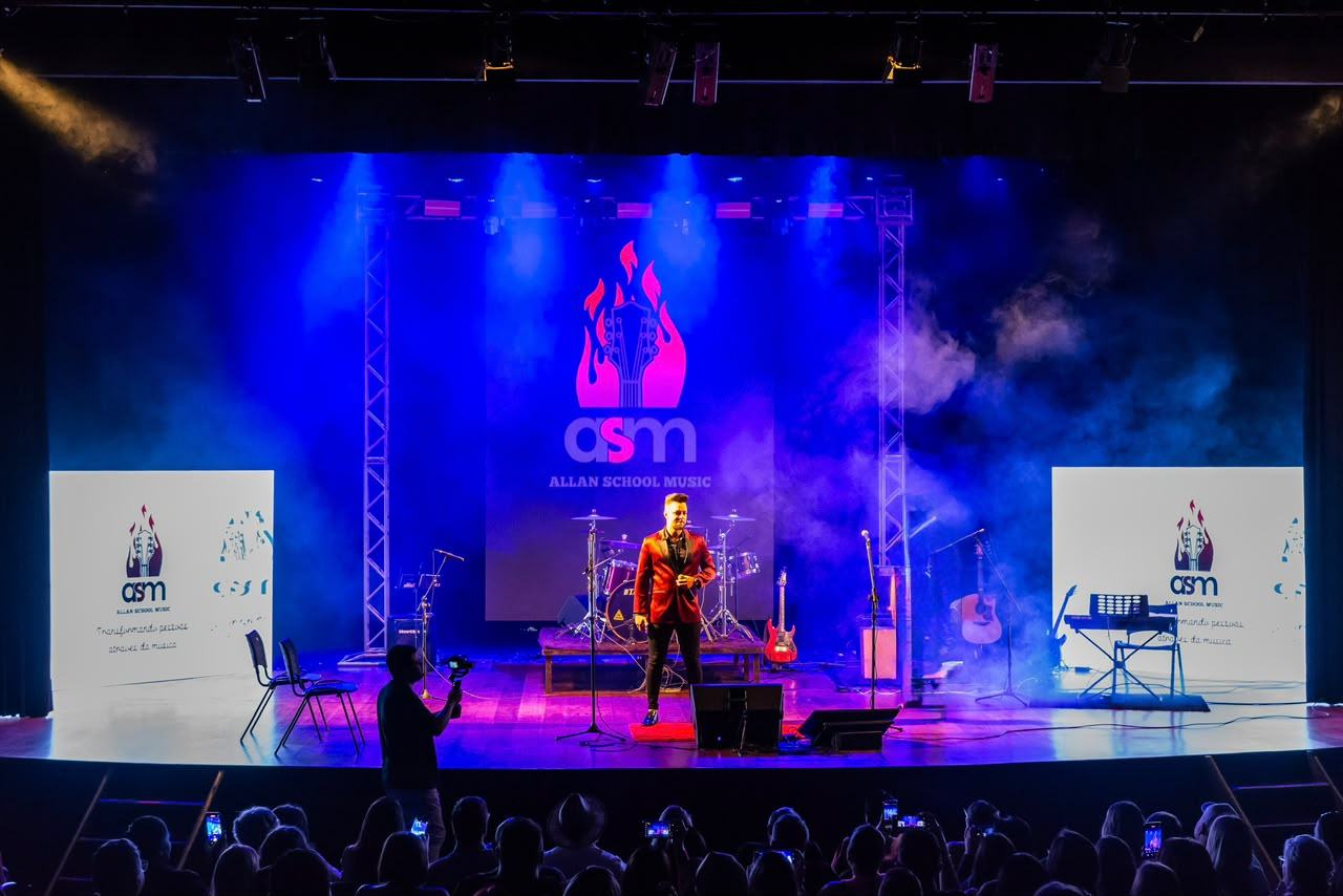 Palco de uma apresentação da ASM com o telão da Allan School Music e o Allan de paletó vermelho diante da plateia